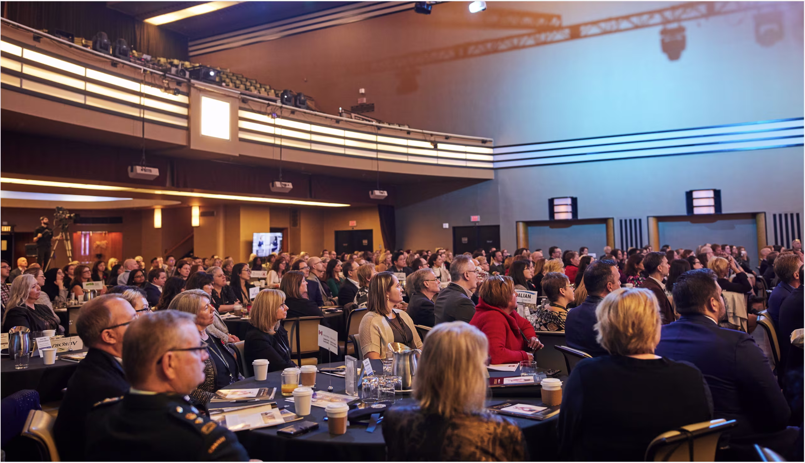 Wide view of a conference hall filled with seated attendees watching a presentation on stage.